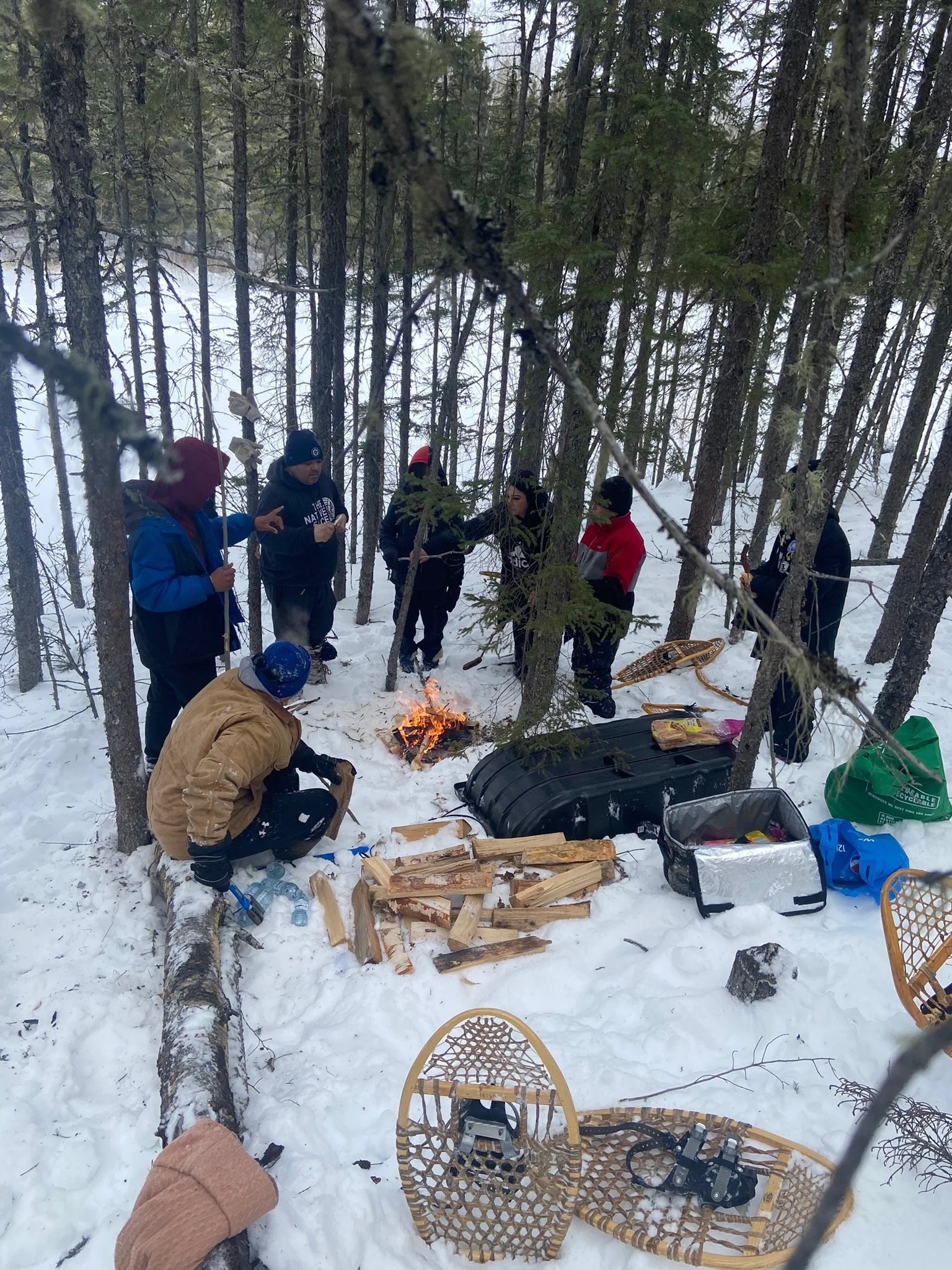 People have gathered in a snowy forest and are gathered around a small campfire, with some chopping wood and others standing around. There are snowshoes and supplies on the snow, and a person kneeling near the fire in a tan jacket and blue helmet.
