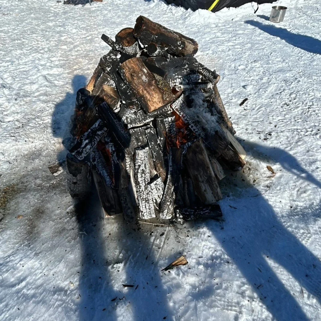 A pile of burning wood logs on snow with smoke rising in cold outdoor setting.