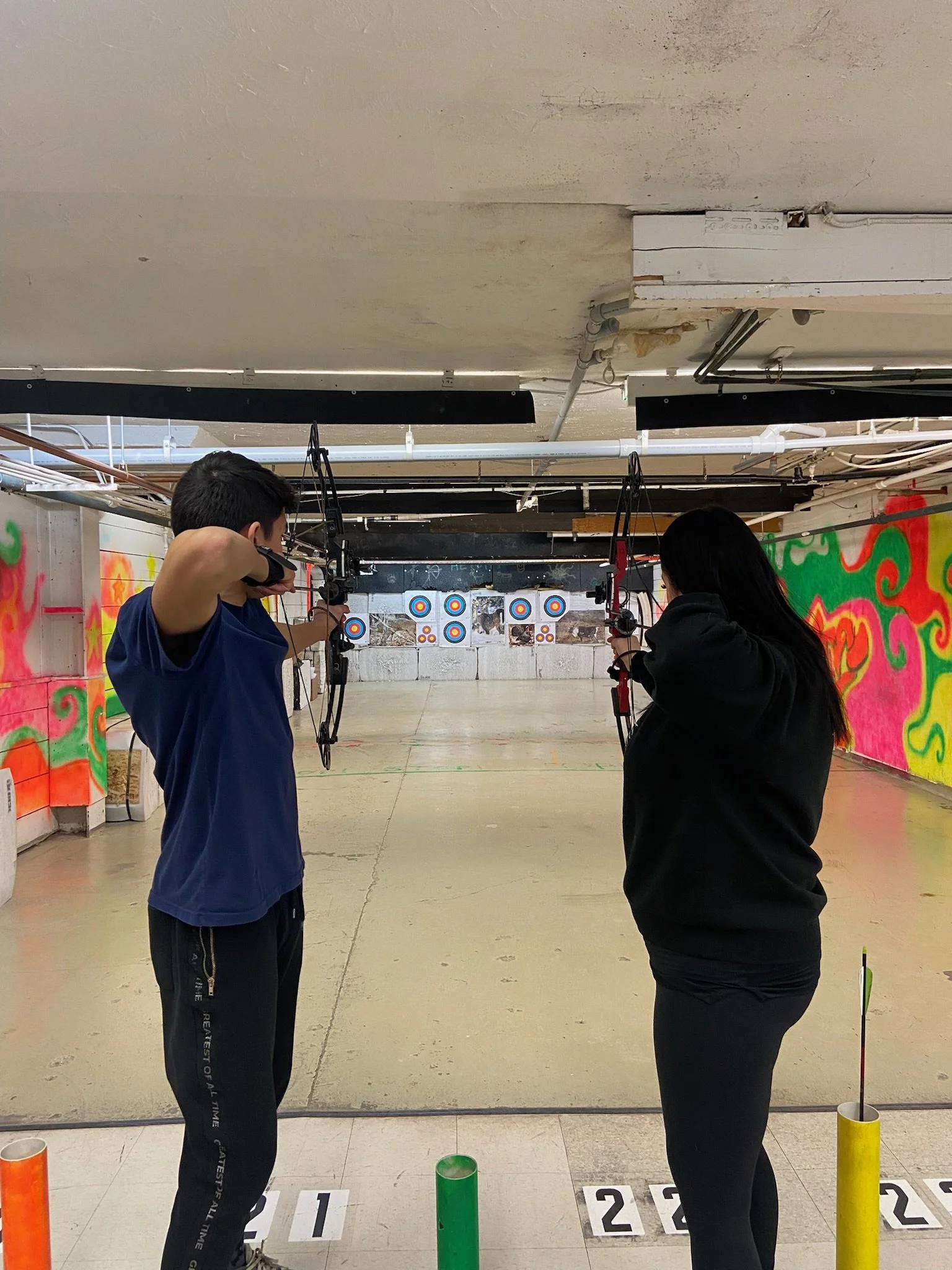 Two individuals aiming bows at archery targets inside an indoor archery range, with colorful wall murals on the sides and numbered lanes on the floor.
