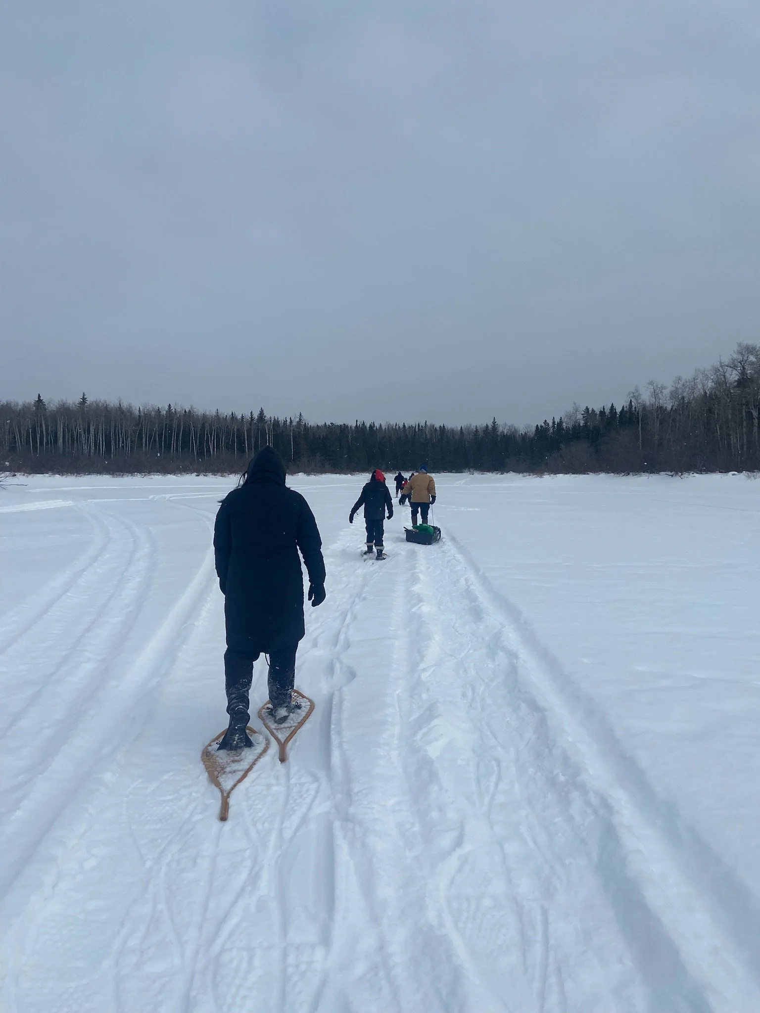 People walking on snow-covered frozen lake with sleds and snowshoes, surrounded by trees under overcast sky