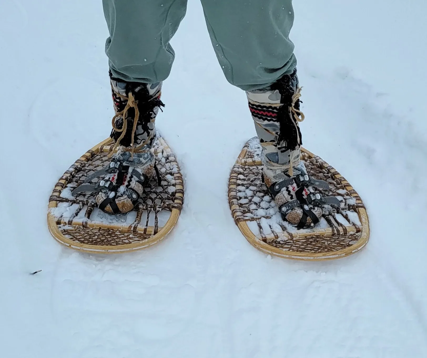 Person standing on snow wearing snowshoes with patterned boots, pants, and green outerwear.