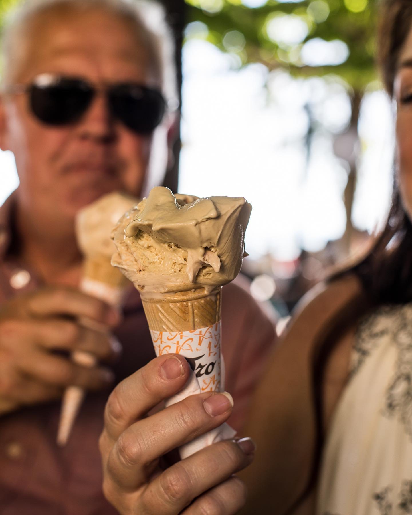 A person holding an ice cream cone topped with chocolate or coffee-flavored ice cream, with two people in the background outdoors.