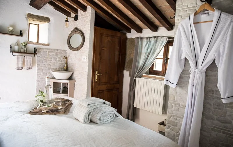 A cozy bedroom with white walls, a bed with folded towels, a robe hanging on the wall, and a bathroom sink near a window with light curtains.