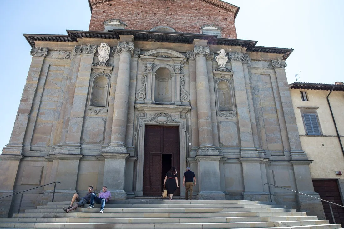 Facade of a historic church with stone columns and decorative architectural details, steps leading up to large wooden doors, and three people walking or sitting nearby.