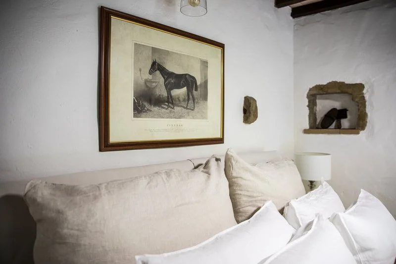A cozy bedroom with a white wall, a framed black-and-white horse illustration, large beige pillows, a white bedside lamp, and a rustic wall shelf with a small decorative item.