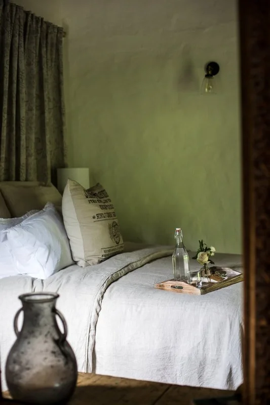 A cozy bedroom with a bed featuring white linens, beige pillows, and a beige duvet. A wooden tray with a glass bottle, a small vase with flowers, and a book is on the bed. There is a vintage glass vase in the foreground and a green wall with a black wall lamp above the bed.