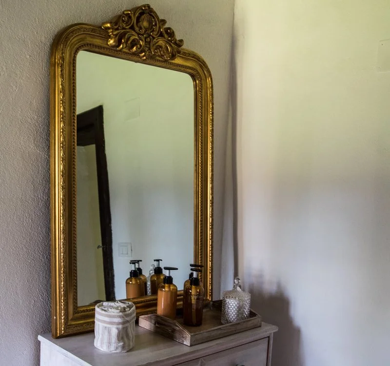 A tall ornate gold-framed mirror reflecting part of a room with a doorway. On a small white wooden dresser in front of the mirror, there are four pump bottles, a ceramic container, a glass jar, and a tray.