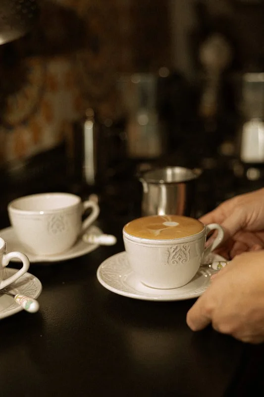 A person preparing to drink coffee in a white teacup with a matching saucer, with another similar teacup and a small milk jug nearby on a black table, in a cozy cafe setting with warm lighting.