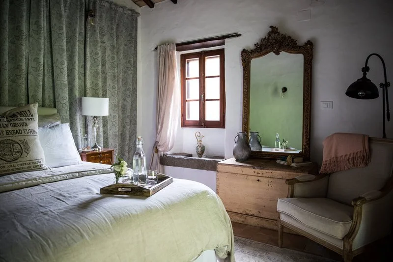 Cozy bedroom with a neatly made bed, white bedding, and a tray with water bottles and a flower. The room has a window with natural light, a vintage mirror with an ornate dark frame, a small chest, a cushioned armchair with a pink throw, and soft neutral colors.