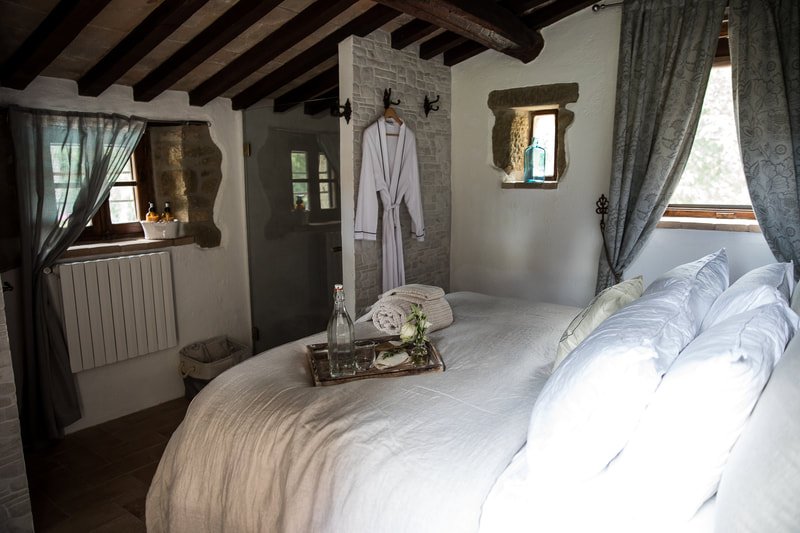 Cozy bedroom with a large bed covered in white linens, pillows, and a tray with water and a towel on top. Two small windows with curtains, a robe hanging on hooks, and rustic wood beamed ceiling. The room has a mix of white walls and stone accents.