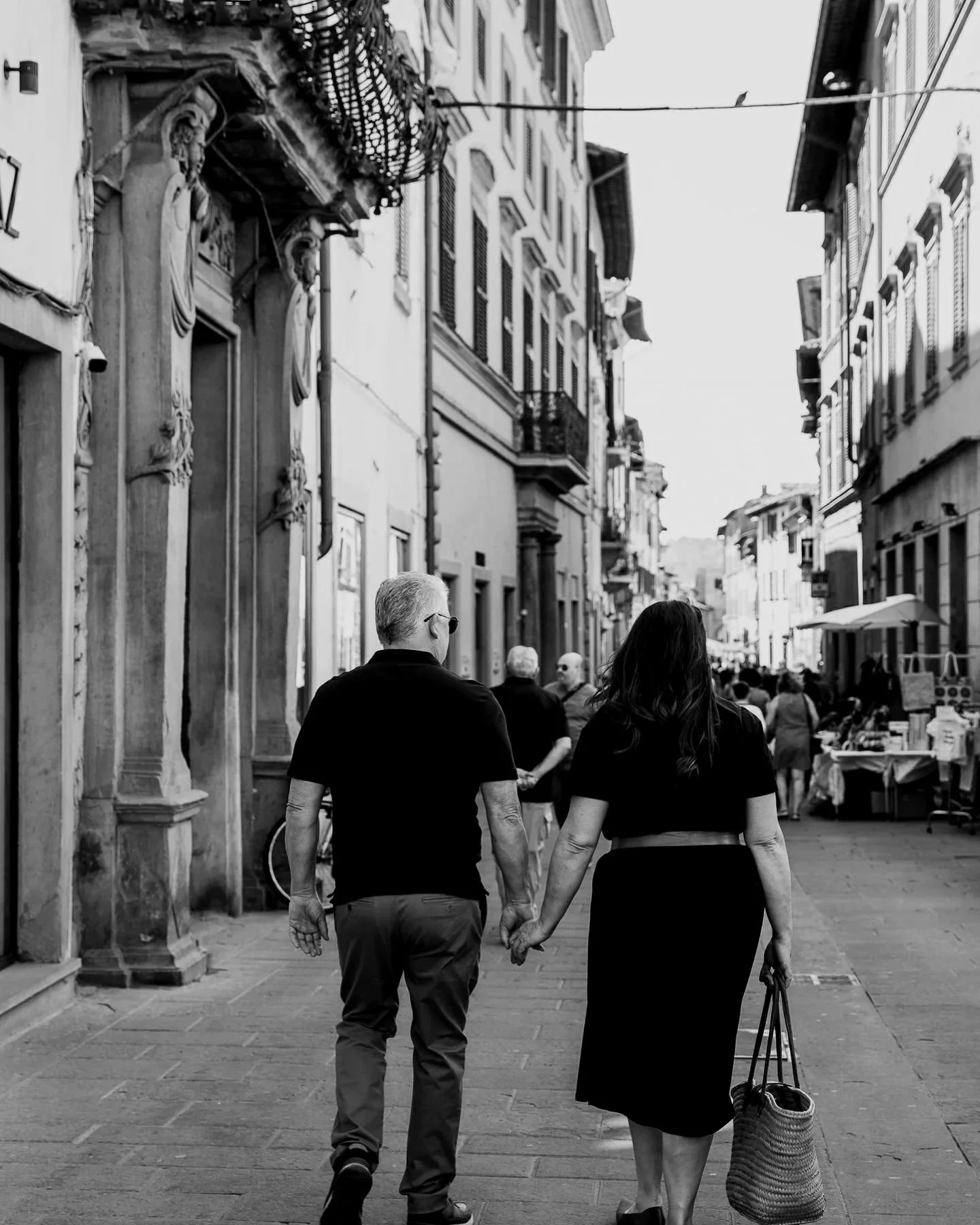 A black-and-white photo of a couple walking hand-in-hand down a crowded street in a European city, with old buildings and shop stalls on the side.