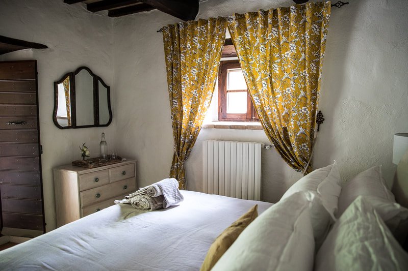 Cozy bedroom with white bed, yellow floral curtains, wooden window, dresser with mirror, small decor, and rustic ceiling beams.