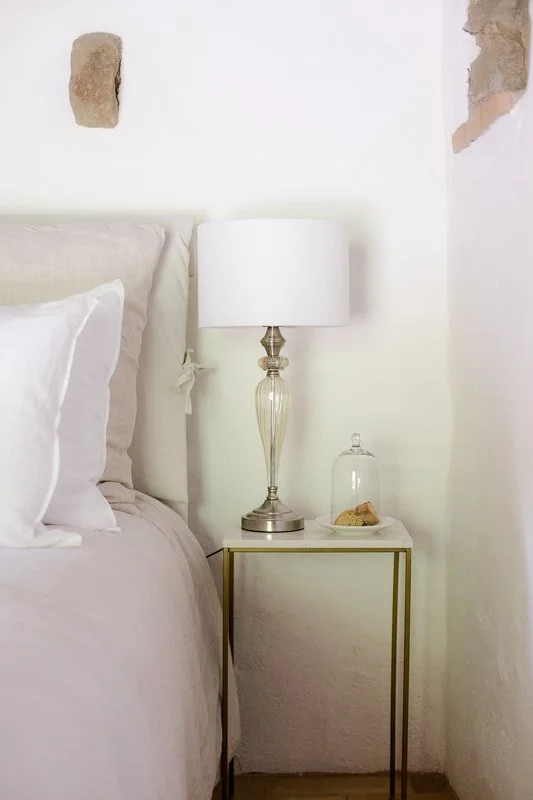A bedroom corner with a white bed, a white nightstand, a tall glass lamp with a white shade, and a glass dome covering a plate with baked goods. The wall behind has exposed bricks in some areas.