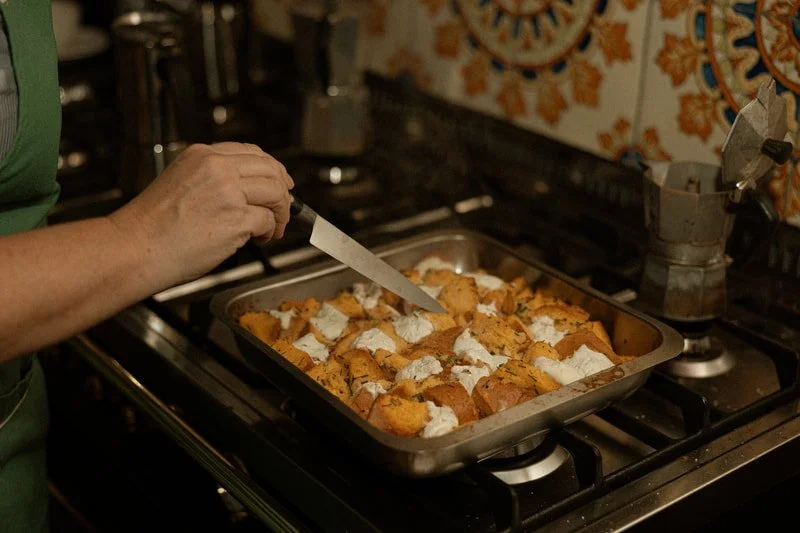 A person adding dollops of white cream or sauce onto a tray of roasted sweet potatoes or squash in a kitchen setting.