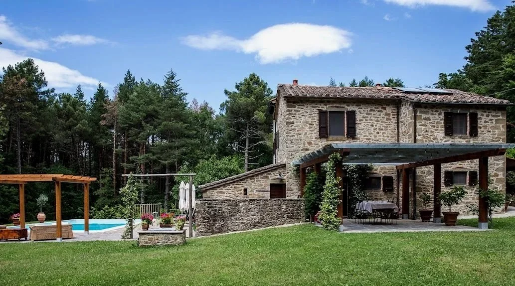 Stone house with patio, lush green lawn, outdoor dining area, garden, and pool in the backyard, surrounded by trees under a partly cloudy sky.