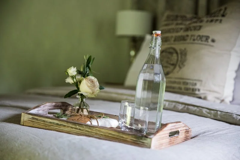 A wooden tray on a bed holding a clear glass water bottle with a swing top, a glass, and a small bouquet of white roses with green leaves, with a beige pillow and part of a lamp in the background.