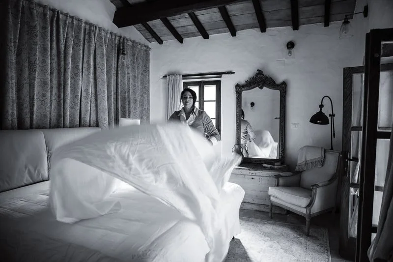 A woman making a bed in a rustic bedroom with wooden ceiling beams, a mirror, and vintage furniture.