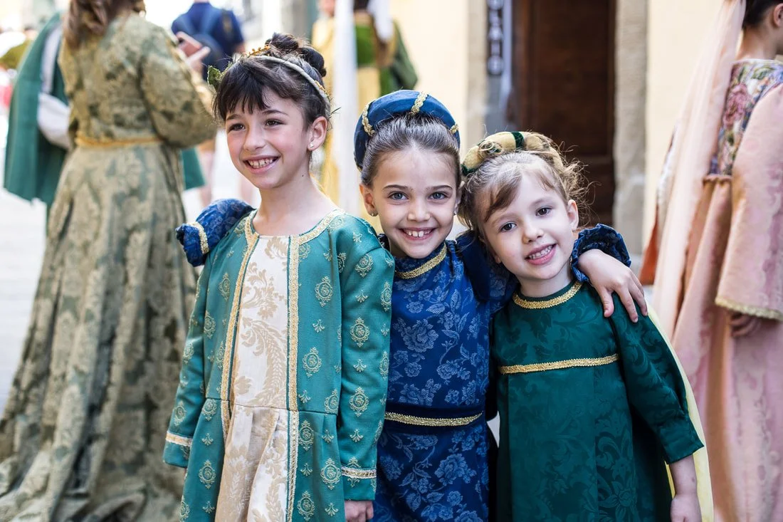Three young girls in colorful, elaborate costumes smiling and posing for a photo at a cultural event or festival.