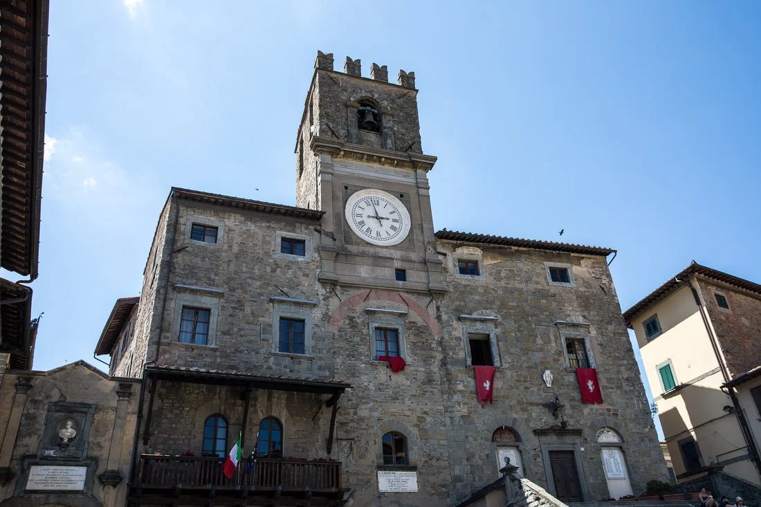 An old stone clock tower with Roman numerals, flags, and banners in a European town square under a clear blue sky.