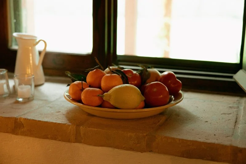 A ceramic bowl filled with oranges, apples, and a lemon sits on a windowsill of a kitchen counter.