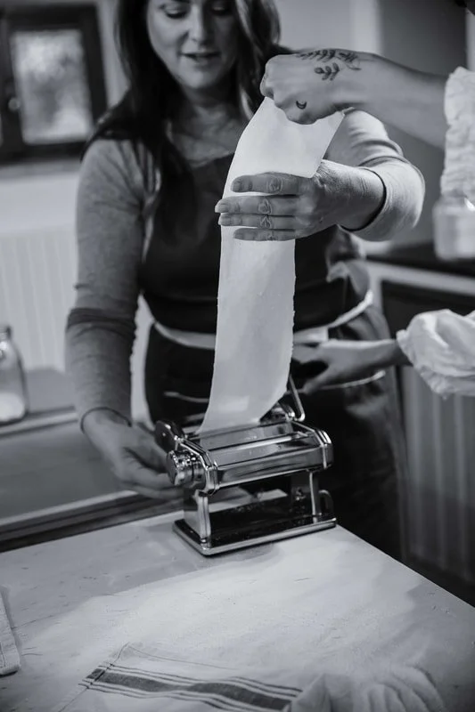 A woman is using a pasta machine to roll out fresh pasta dough in a kitchen.