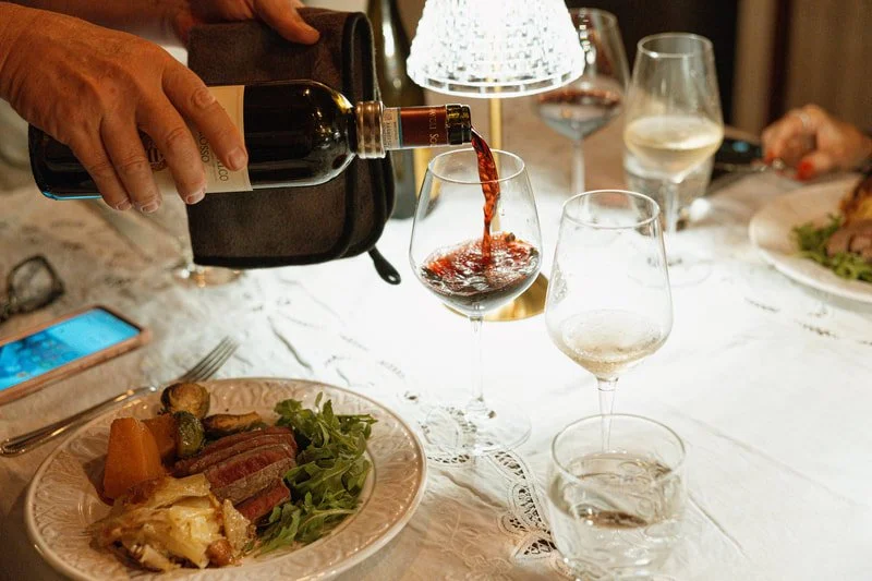 Person pouring red wine into a glass on a dining table with plates of food and glasses of water, illuminated by a decorative lamp.
