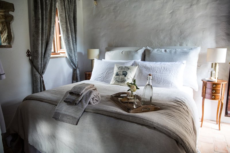 Cozy bedroom with white bedding, decorative pillows, wooden nightstands, table lamps, and a tray with glass water bottles and flowers.