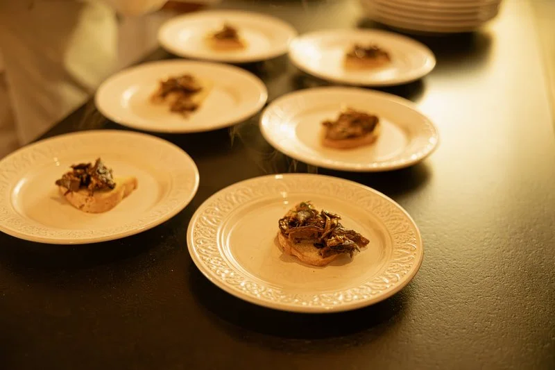 Six small white plates with food, arranged on a dark table, with a dark background.