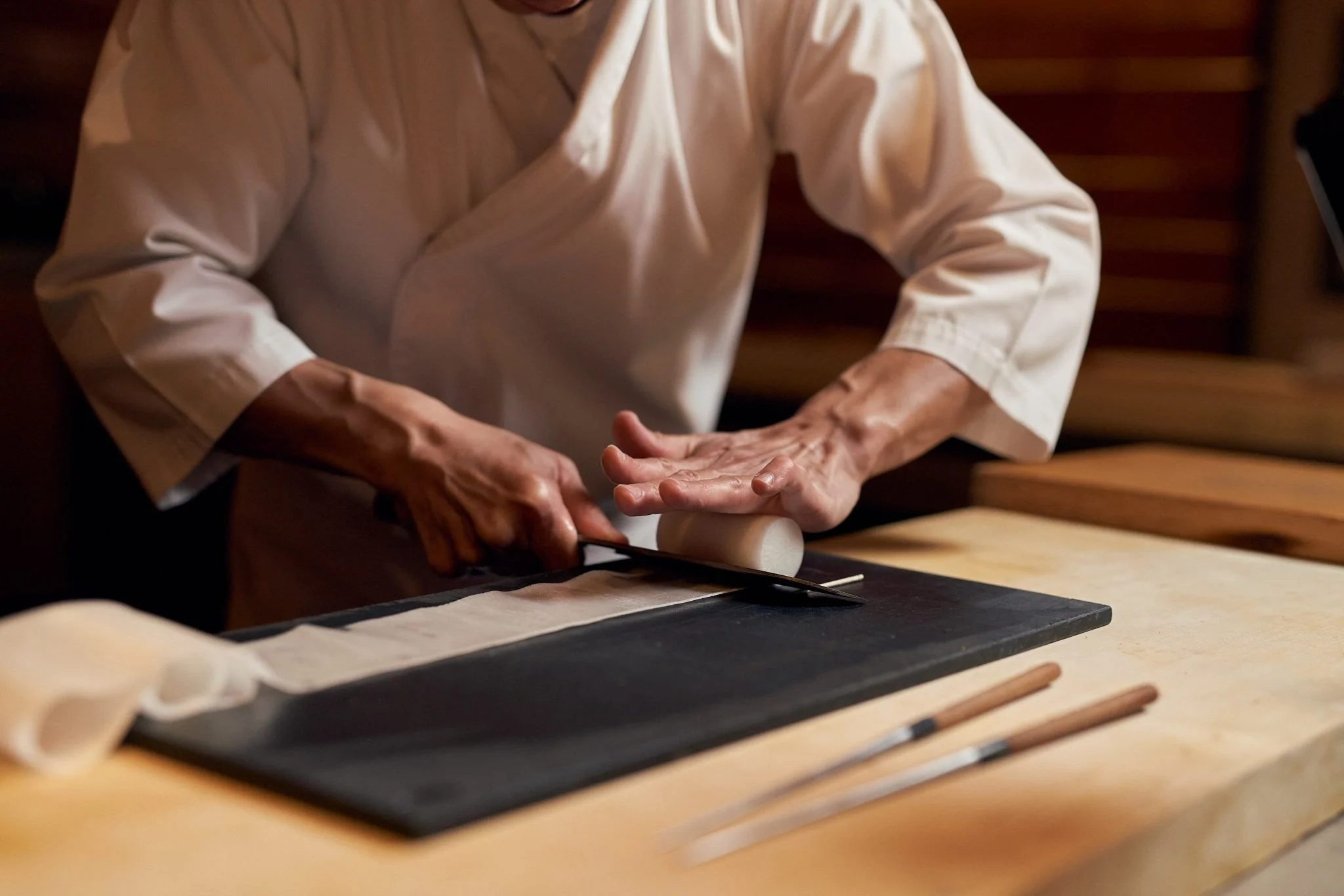 A person in a white chef's coat slices a roll of dough with a knife on a black cutting board in a kitchen.