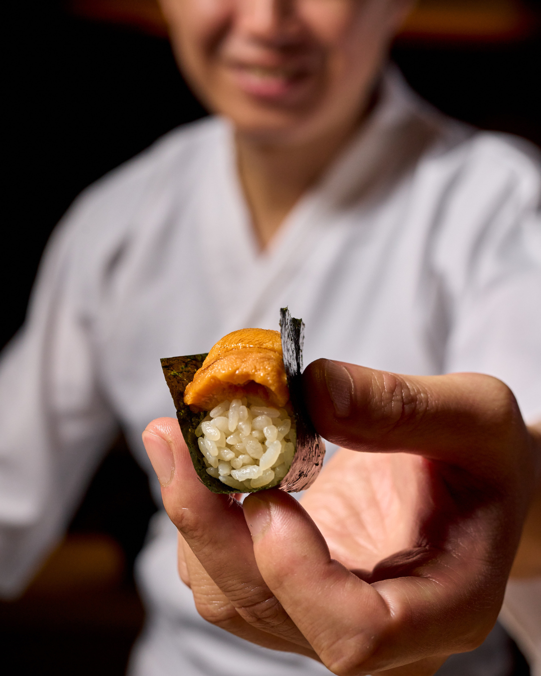 The Den Miami close-up of a sushi roll with rice and salmon, held by black chopsticks, with other sushi rolls on a white plate and traditional Japanese dishes in the background.