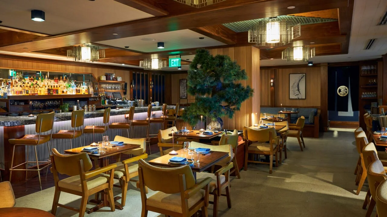 A cozy restaurant interior with wooden walls and ceiling, a bar with bottles of alcohol, a large green tree in the center, and neatly set tables with blue napkins and glassware.