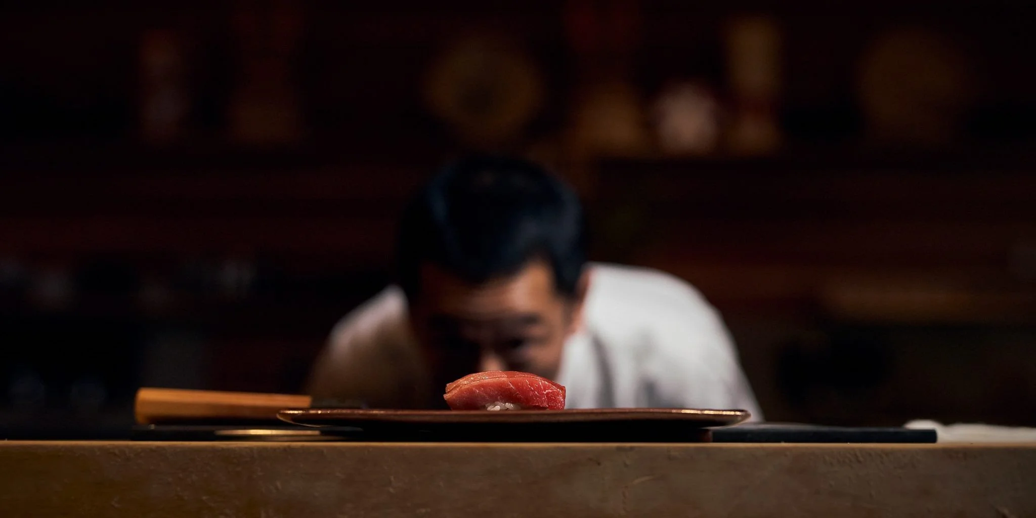 A chef looking closely at a piece of sushi on a plate in a dimly lit setting.