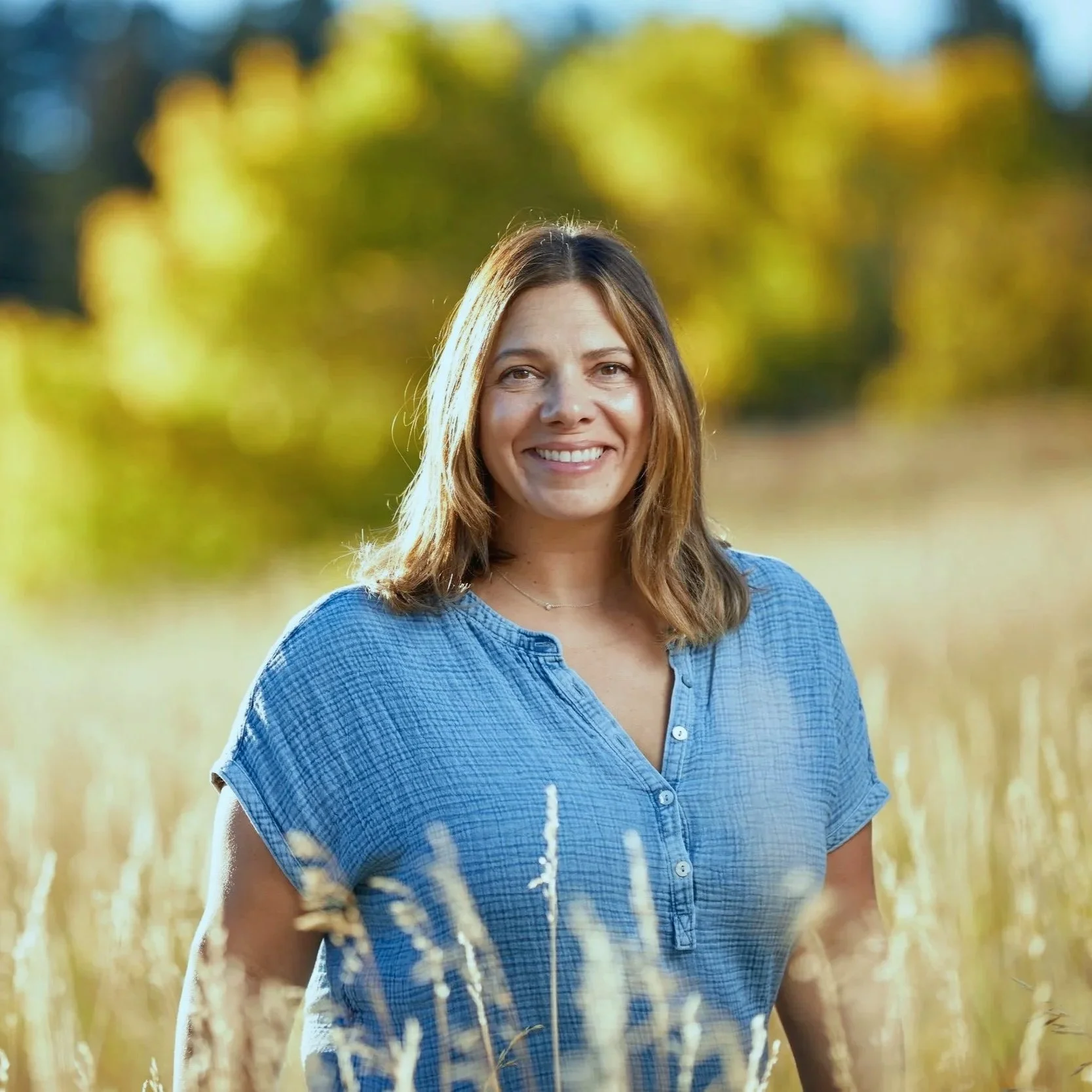 A woman smiling outdoors in a field of tall grass with trees in the background feeling calm and restored.