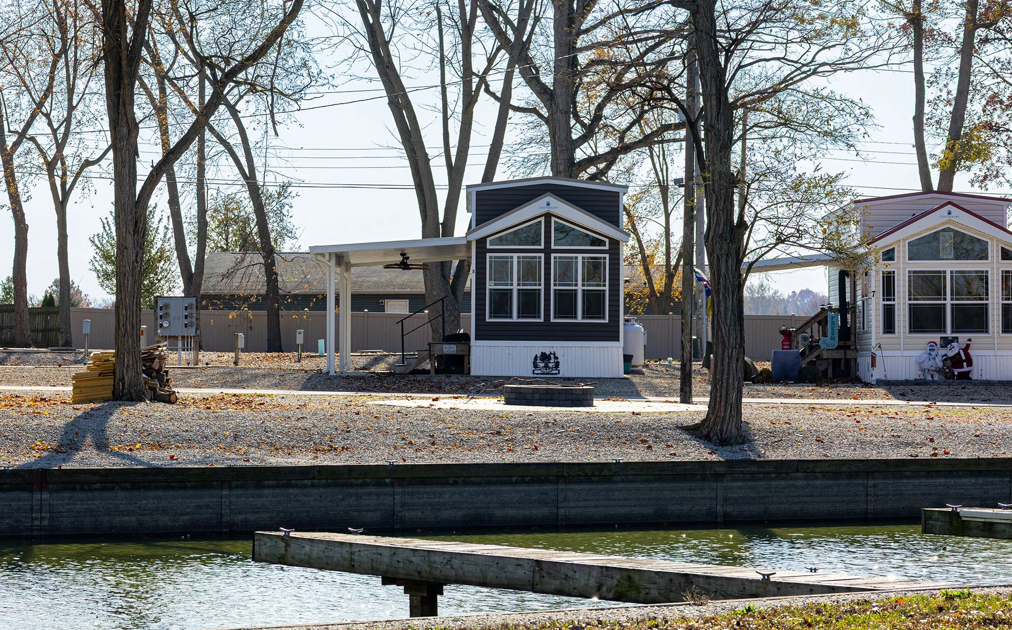 View of houses along a canal with trees and fallen leaves, one house painted dark blue with large windows, and another house painted white with Christmas decorations.