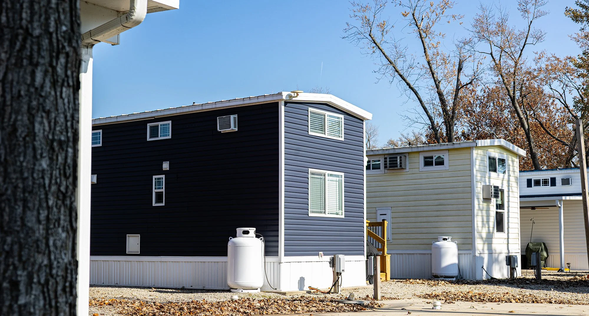 A row of small, colorful manufactured homes with metal siding, some with air conditioning units in the windows, set against a background of trees with autumn leaves and a clear blue sky.