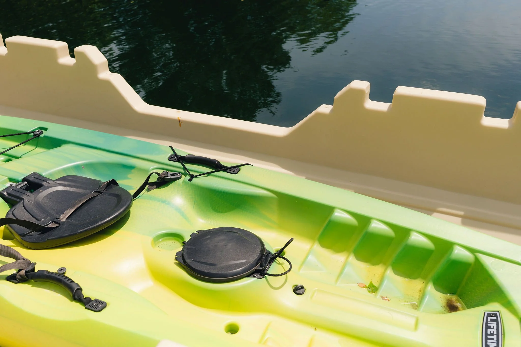 Close-up of a green kayak with black accessories floating on water, with a beige barrier along the edge of the water.