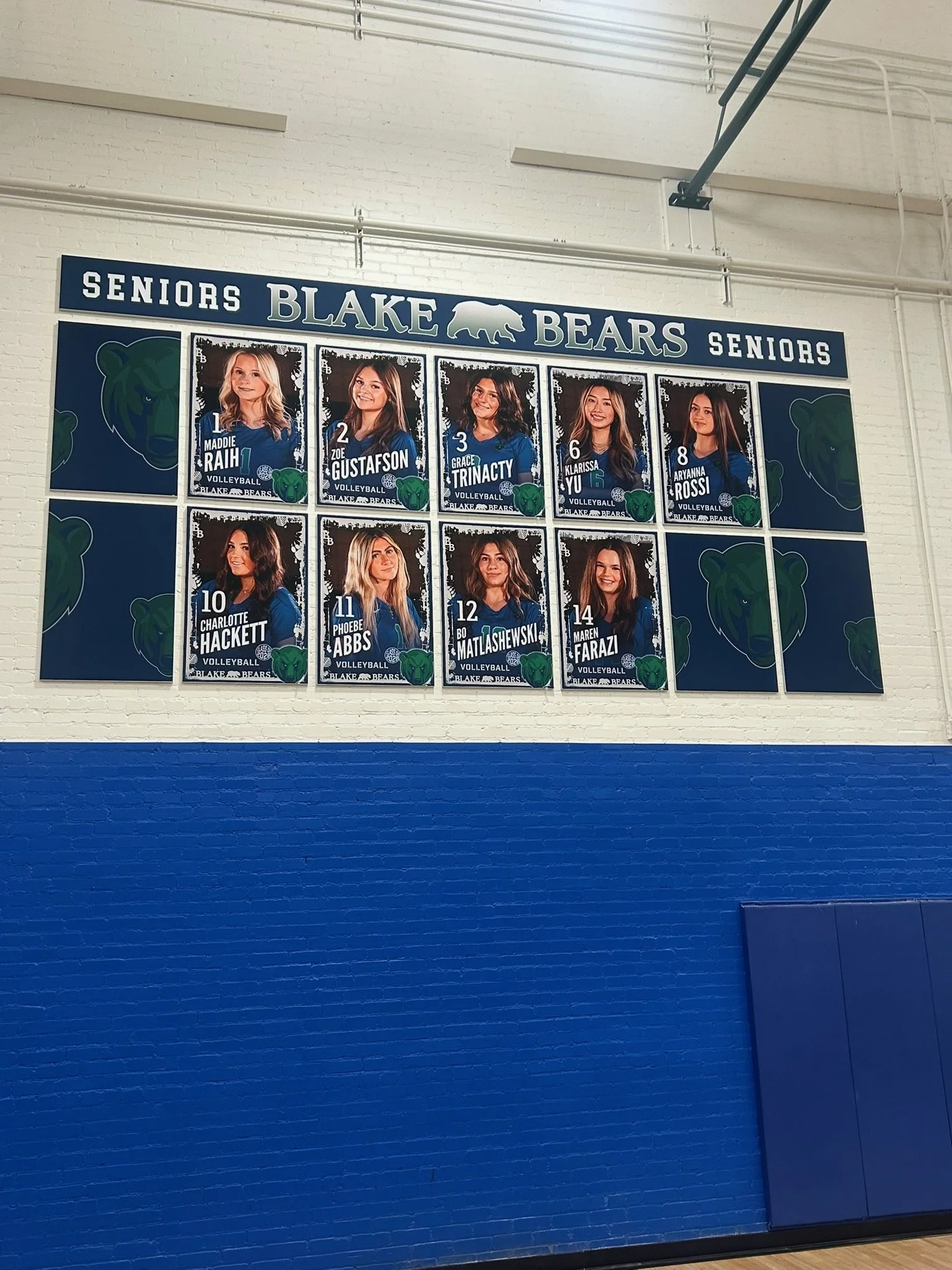 School gymnasium wall display featuring senior volleyball player posters for Blake Bears, arranged in a grid. Posters include photos and names of players Maddie Rahi, Zoe Gustafson, Grace Trinity, Klarissa Yu, Arianna Rossi, Charlotte Hackett, Phoebe
