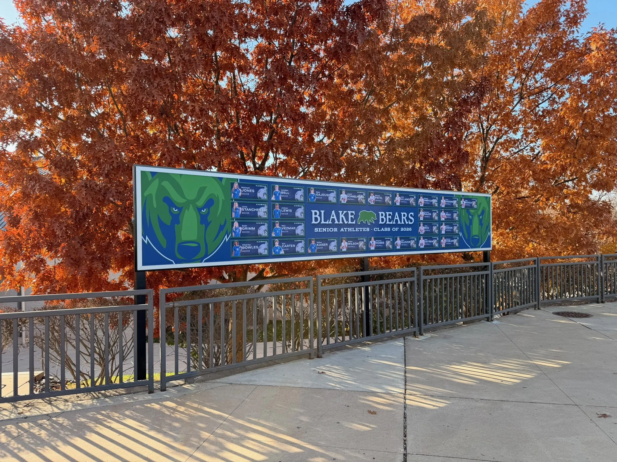A billboard honoring Blake Bears senior athletes from the class of 2026, with a large bear logo and individual photos of athletes, located outdoors in front of autumn-colored trees and a metal railing.