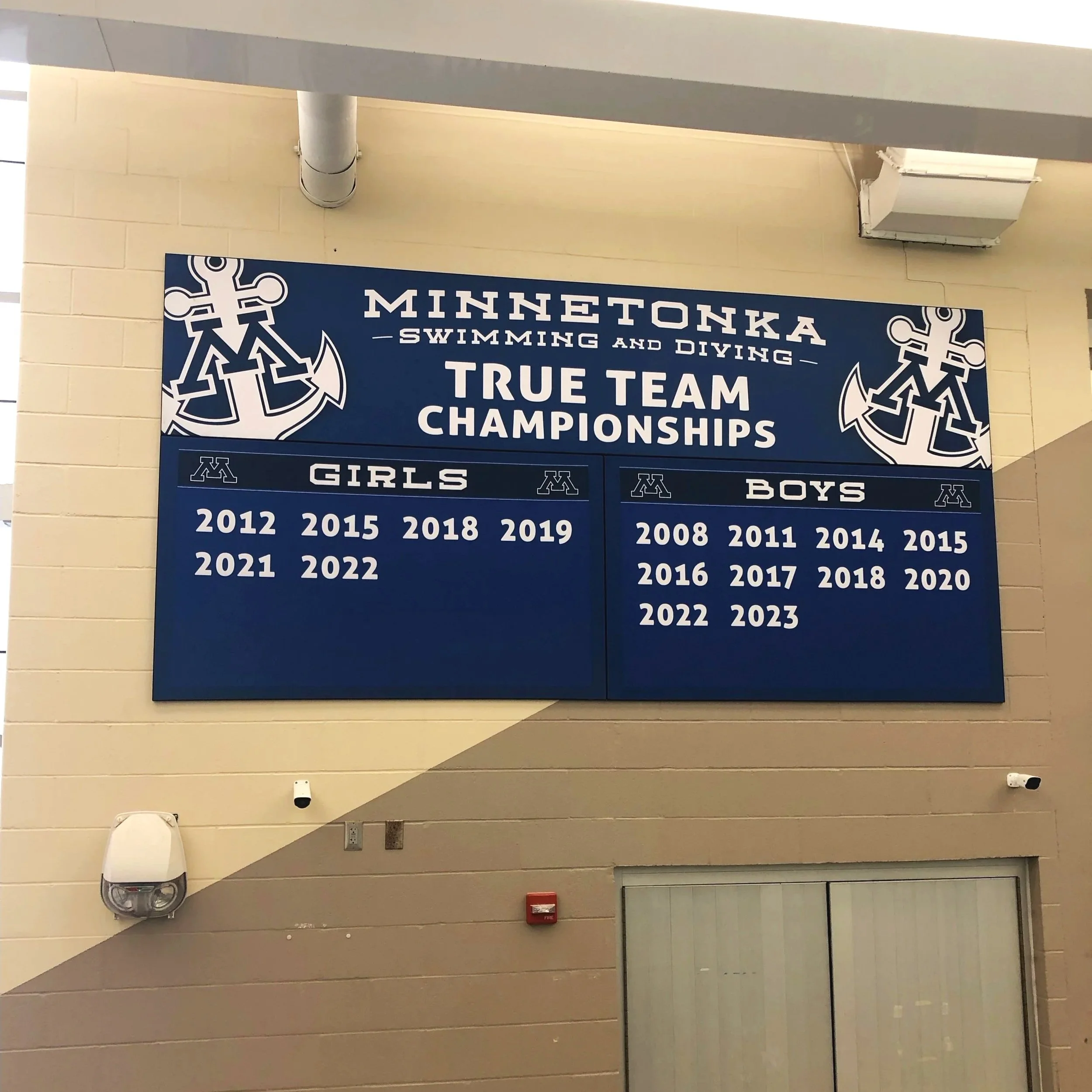 A scoreboard at Minnetonka Swimming and Diving True Team Championships listing winners for Girls and Boys teams with years from 2012 to 2023.