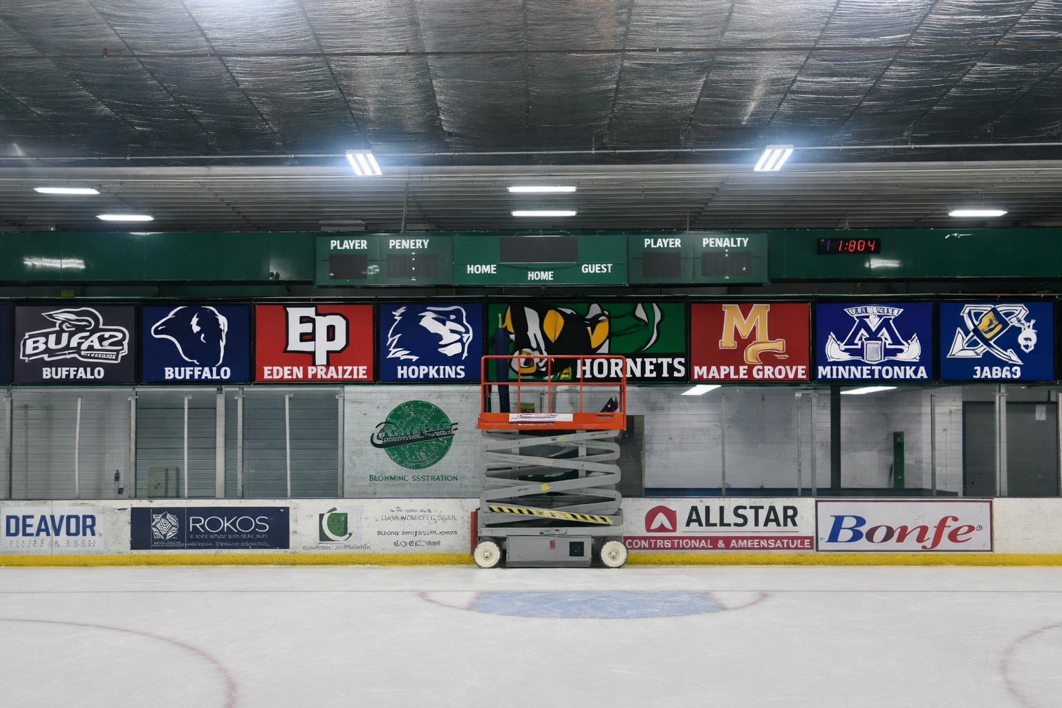 Empty ice hockey rink with a scissor lift in front of team banners and advertising. The banners display logos and names of local teams, including Buffalo, Eden Prairies, Hopkins, Hornets, Maple Grove, Minnetonka, and JAB6. The scoreboard is visible a