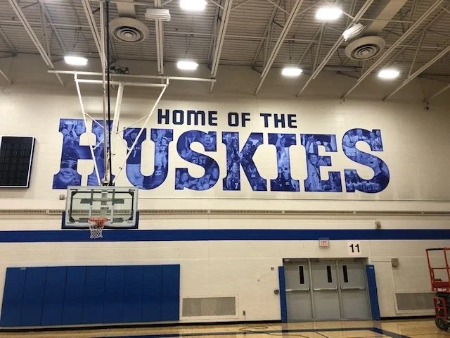 Gymnasium wall with a large sign that says 'HOME OF THE HUSKIES' with a basketball hoop in front.