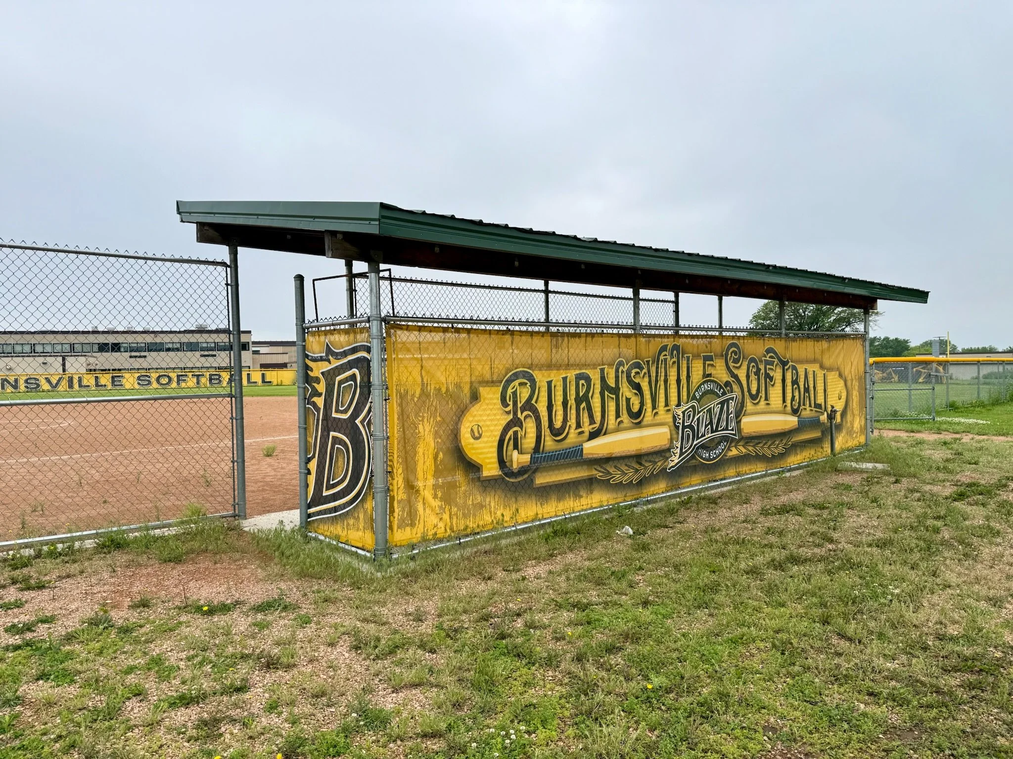 Fenced softball field with a yellow banner that reads 'BURNSVILLE SOFTBALL' and 'BURNSVILLE HIGH SCHOOL'. Dugout with a green roof and a bench underneath.
