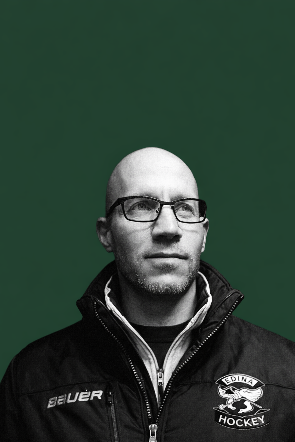 A bald man with glasses and a goatee, wearing a black Edina Hockey jacket, looking upwards against a green background.