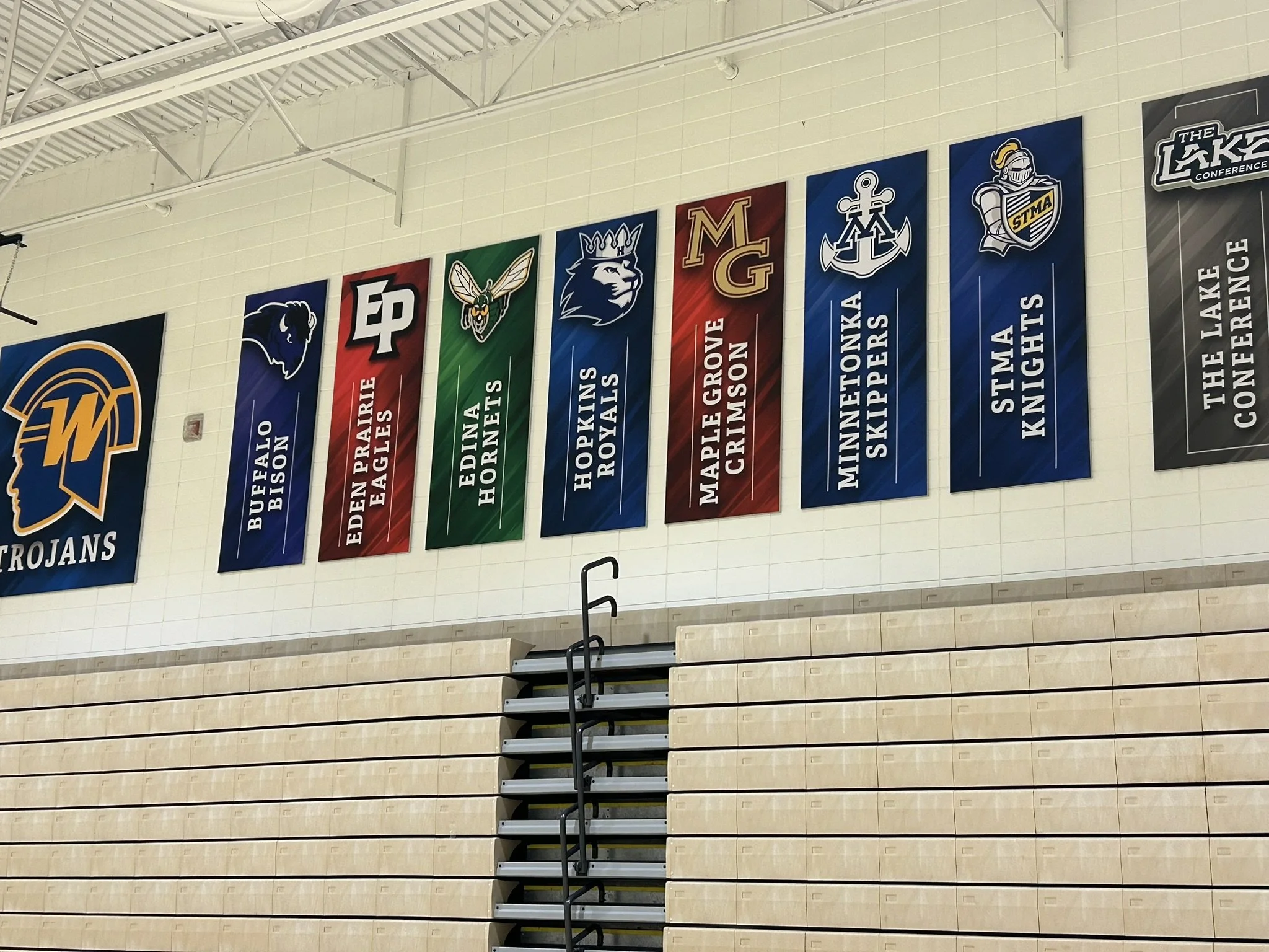 Banners of different high school team logos and names hanging on a gymnasium wall, including Buffalo Bison, Eden Prairie Eagles, Edina Hornets, Hopkins Royals, Maple Grove Crimson, Minnetonka Skippers, STMA Knights, and The Lake Conference logo.