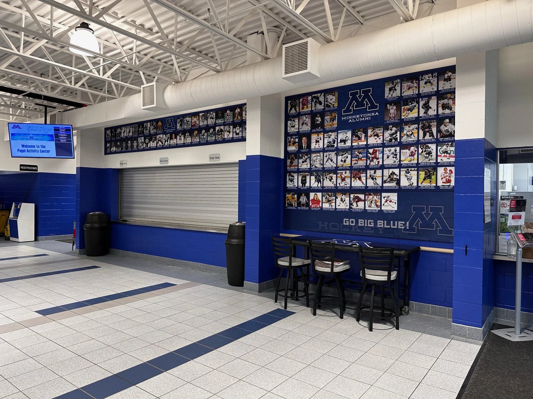 Indoor view of the Minotonka Arena, showing a wall with numerous hockey player photographs, a closed concession window, a seating area with three chairs, and a digital display sign.