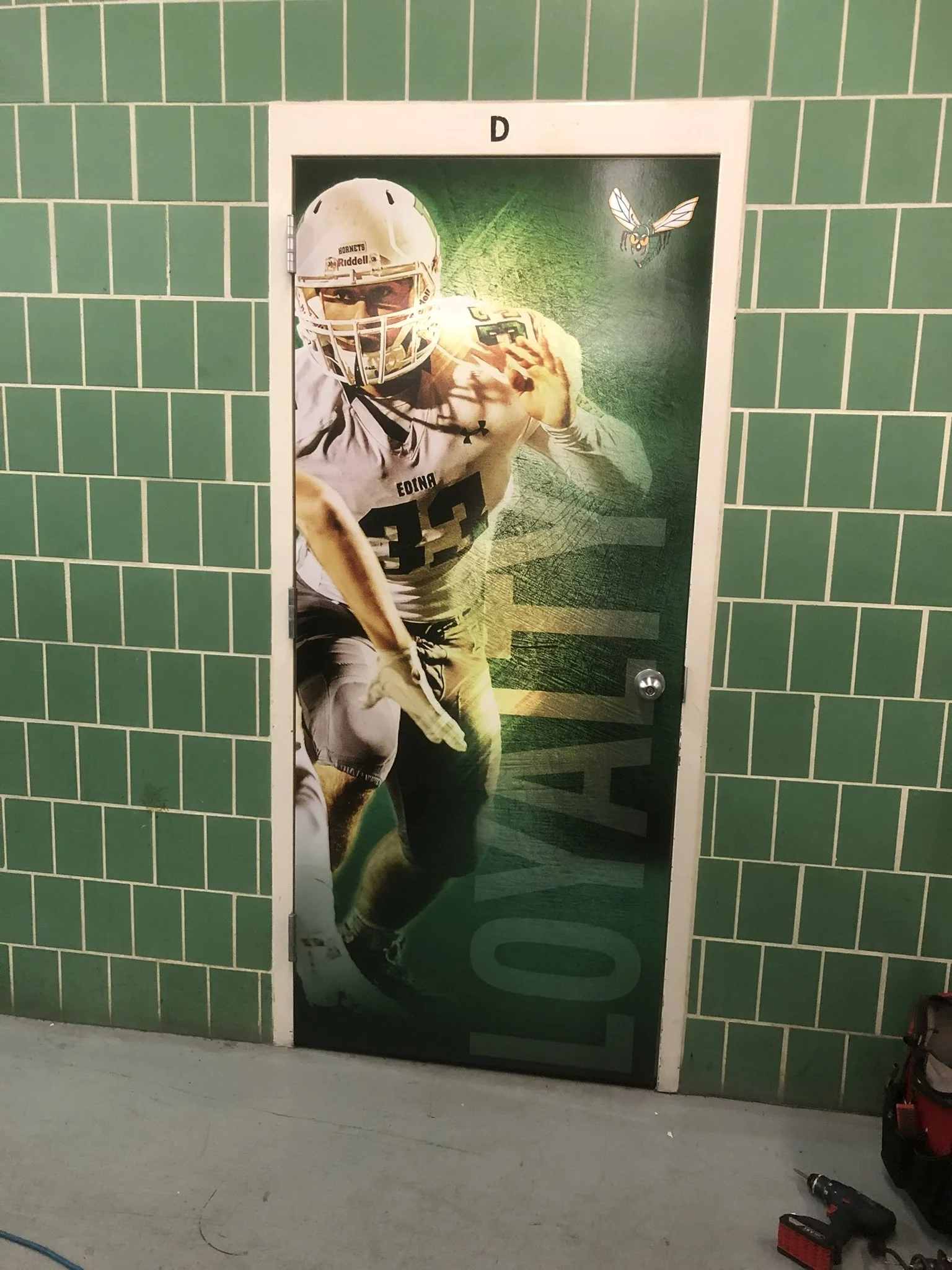 Door with a large sports poster showing a football player in uniform running and holding a football, with a hornet logo in the top right corner and green tiled walls around.