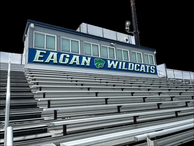 View of empty bleacher seats under the press box at Eagan High School stadium with a sign that reads 'Eagan Wildcats' and a stylized wildcat logo, against a black sky.