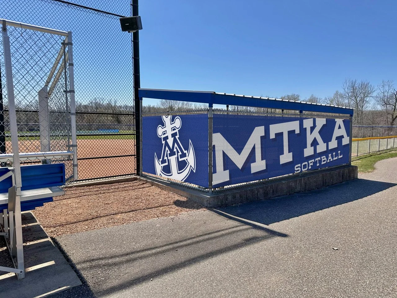 View of a softball field with a blue banner that reads 'MTKA SOFTBALL' and features an anchor and cross design. The field has a chain-link fence, a backstop, and a clear blue sky.
