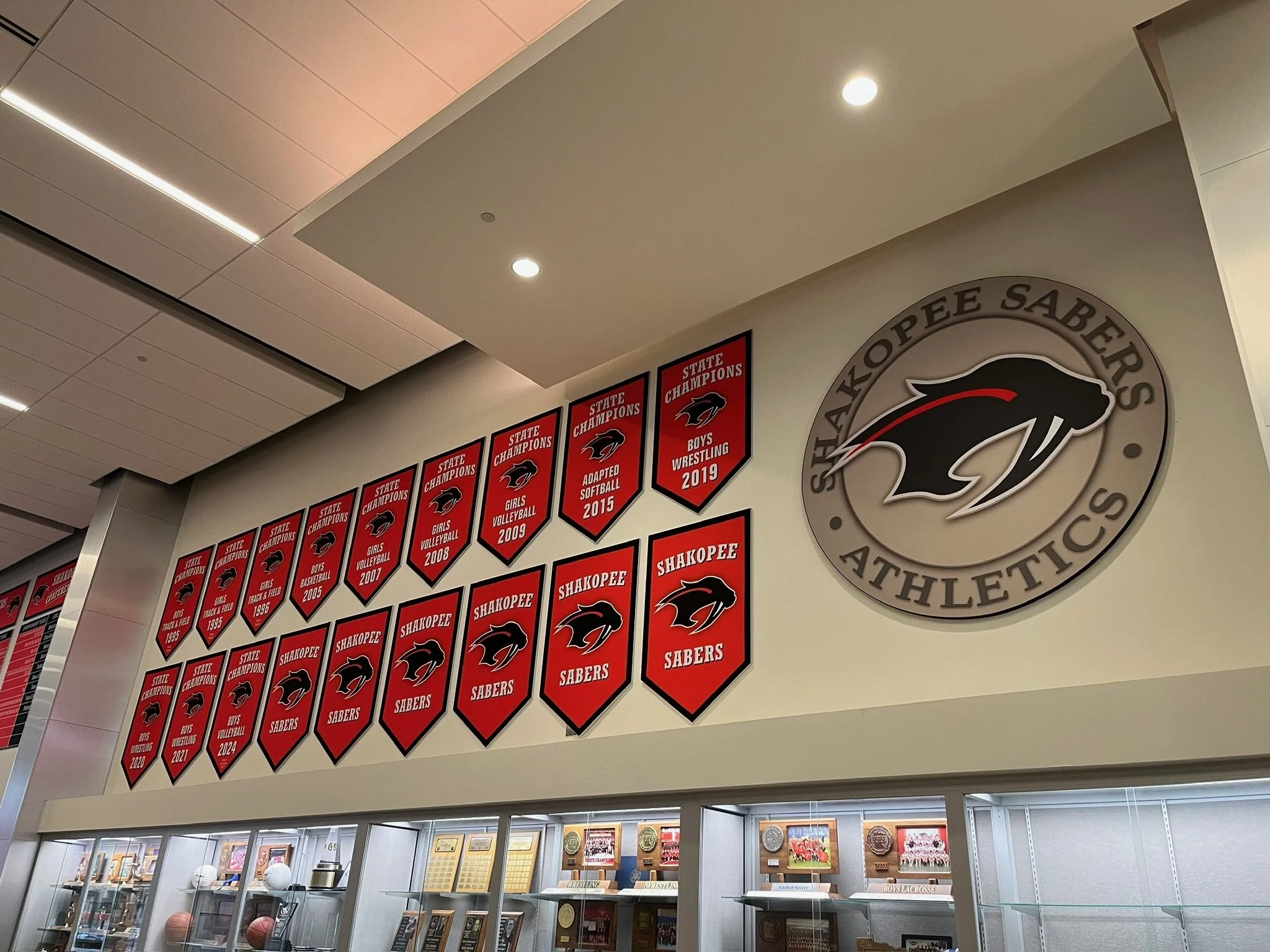 A wall display in a sports facility featuring championship banners for the Shakopee Sabers with many banners for various sports and years, along with a large circular logo for Shakopee Sabers Athletics showing a saber-toothed cat head.
