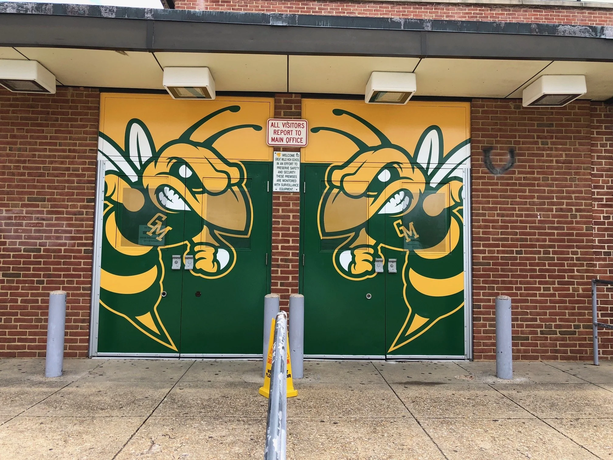 Double doors with green and yellow graffiti of a hornet mascot wearing a bee suit with the letters 'GM' on its chest, set in a brick building exterior. A sign above the doors reads 'All Visitors Report to Main Office' with additional instructions bel